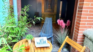 Una mesa de madera con una taza de té, una silla y un banco frente a un edificio con pared de ladrillo y suelo de baldosas.