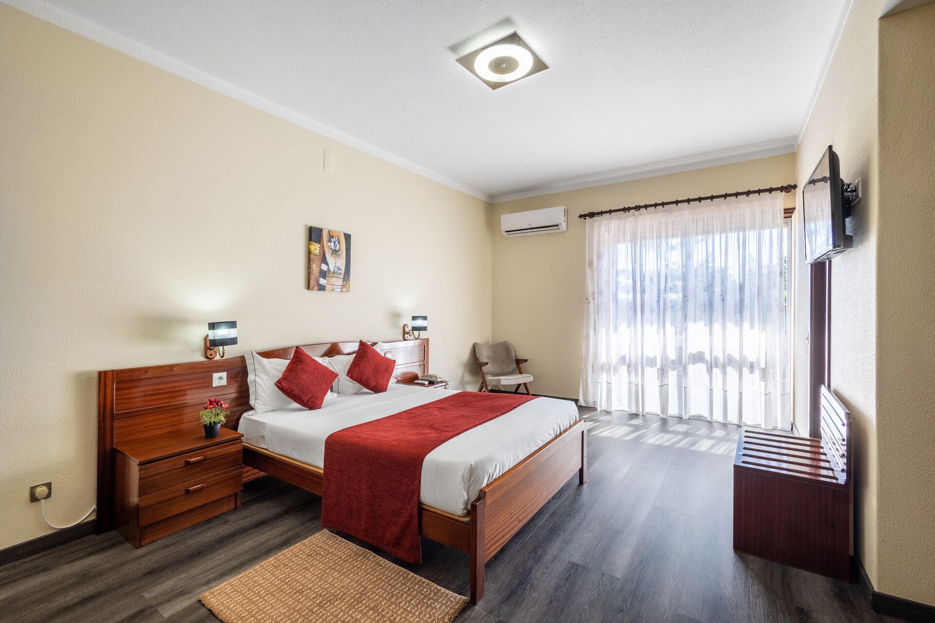 A hotel room with a wooden bed, a red throw blanket, a wooden nightstand, a small table with a lamp, a chair, a TV mounted on the wall, and a window with curtains.