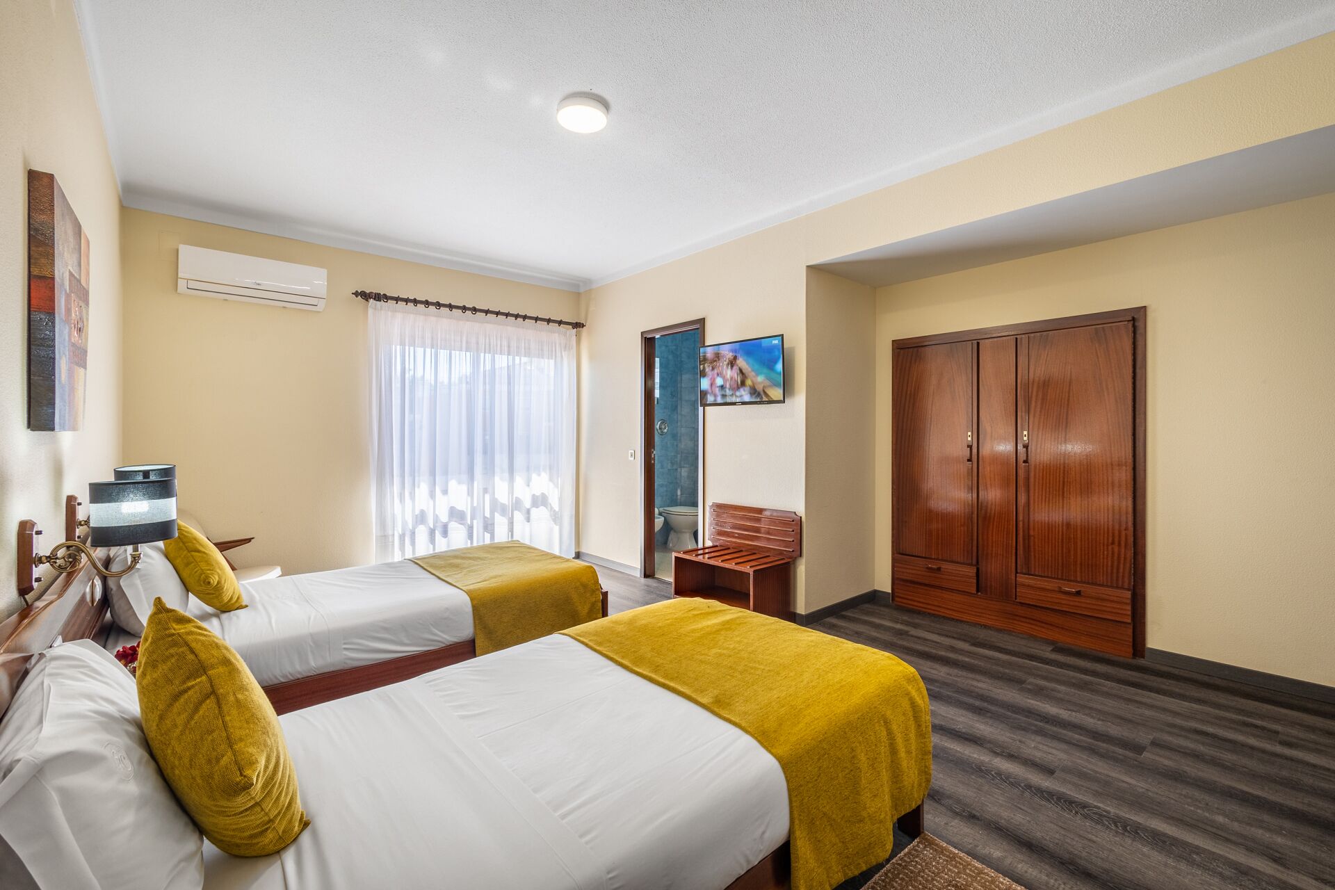 A hotel room with two beds, a wooden wardrobe, a television, and a bathroom visible through an open door.