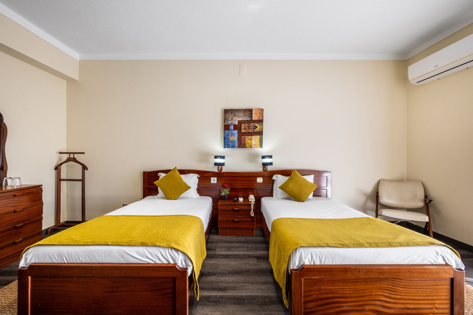 A hotel room with two single beds, a wooden dresser, a chair, and a wall-mounted air conditioner.