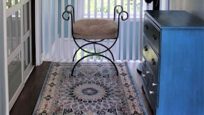 Una habitación con una pared azul, un ventanal con marco blanco, un tapiz estampado, una silla, un aparador y una lámpara colgante.