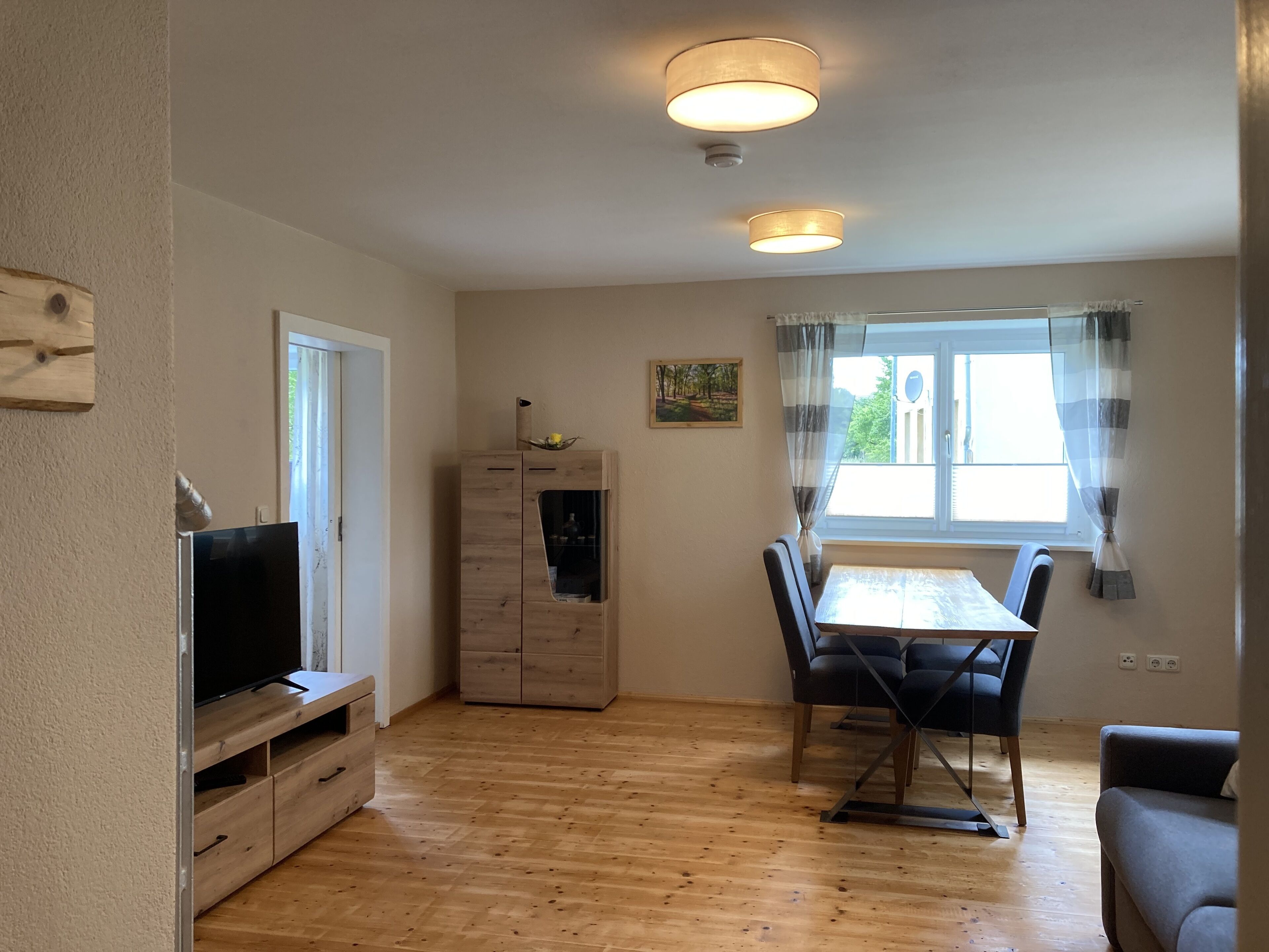 A living room with a TV, wooden cabinet, dining table with chairs, and a window with curtains.