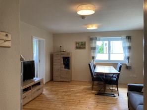 A living room with a TV, wooden cabinet, dining table with chairs, and a window with curtains.