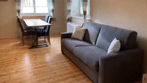 Un salon avec un canapé gris, une table à manger en bois avec des chaises et une fenêtre avec des rideaux.