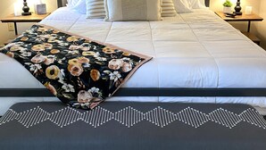 Ein ordentlich gemachtes Bett mit weißer Bettwäsche und einem floralen Überwurf, begleitet von zwei Nachttischen mit Lampen und einer kleinen Pflanze.