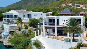 A coastal property with a cliffside pool, multiple-story buildings, and a mountain backdrop. - Seafront Villa in Lagun Curaçao -pool-Wifi + With Private access to sea! (Lagun)