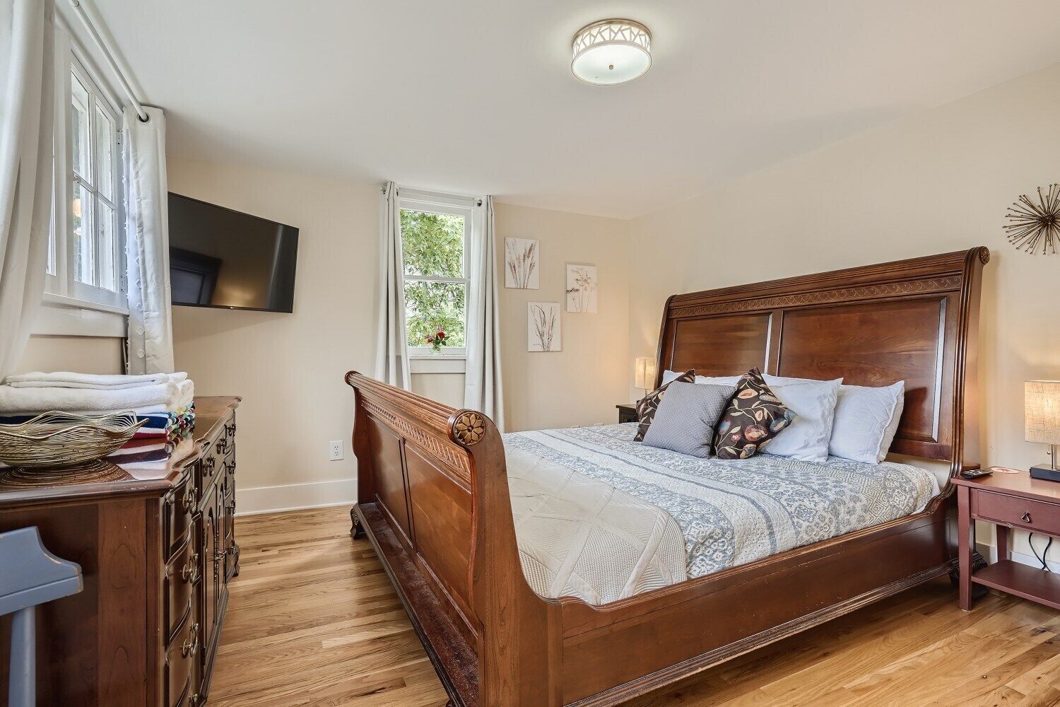 A bedroom with a wooden bed, a dresser, a television on the wall, and a window with curtains.