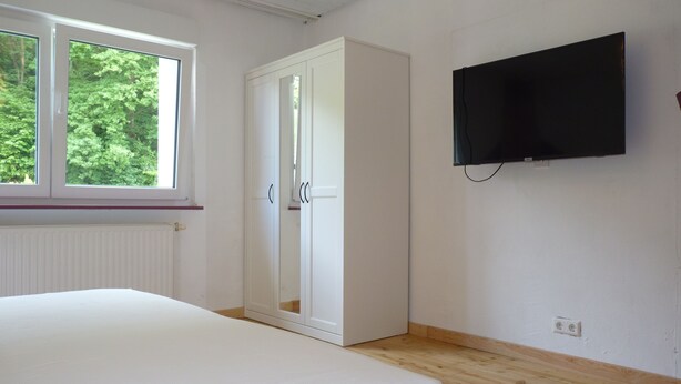 A bedroom with a bed, a wardrobe, a television, and a window with a view of trees.