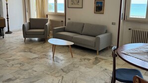 Ein Wohnzimmer mit einer Couch, einem Sessel und einem runden Tisch. Es gibt Fenster mit Vorhängen, eine Heizung und ein Marmorboden.