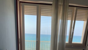 Ein Schlafzimmer mit einem großen Bett, Meerblick durch einen Balkon und einem Fenster mit Jalousien.