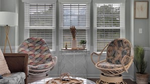 A room with three white-framed windows, wicker chairs with patterned cushions, a small table with a vase and plant, and a lamp.