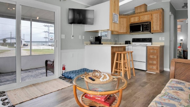 A compact kitchen with wooden cabinets, a microwave, and a stove. There's a glass table with a woven placemat and a chair.