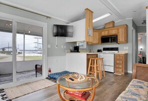 A compact kitchen with wooden cabinets, a microwave, and a stove. There's a glass table with a woven placemat and a chair. - Model Home near Camp Lejeune with Water View (Sneads Ferry)