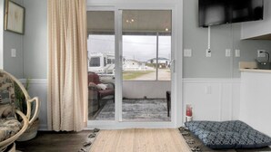 A room with a sliding glass door leading to a covered outdoor area, a television mounted on the wall, and a patterned cushion on the floor.