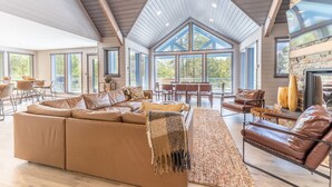Amplia sala de estar con un sofá modular grande, una chimenea y un comedor con techos altos y ventanas amplias.