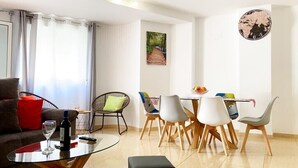 Een moderne woonkamer met een glazen bijzettafeltje, een bank en een eethoek met witte stoelen en een houten tafel.