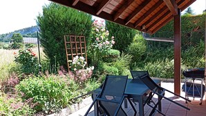 Un patio exterior con una pérgola de madera, una mesa con sillas y un jardín con diversas plantas y flores.
