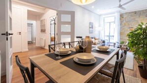 A dining area with a wooden table set for four, black placemats, and a vase. There is a ceiling fan and a large window with a view of the outdoors. - San Martin II | The Rentals Collection (San Sebastián)