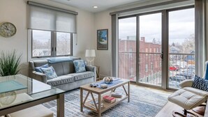 A spacious living room with a large window, a grey sofa, a coffee table, and a glass dining table.
