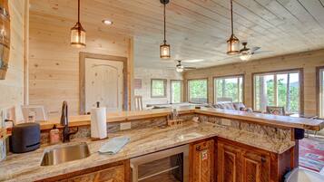 A spacious kitchen with granite countertops, wooden cabinets, and a large island. There are pendant lights, a ceiling fan, and large windows offering views of the outdoors.