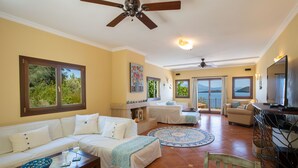 A spacious living area with a sofa, a dining table, and a TV. There are large windows and a ceiling fan. - Private waterfront villa with pool and two private beaches (geni)