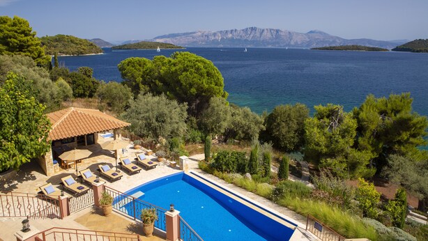 A pool area with lounge chairs and a small pavilion, surrounded by greenery and overlooking a body of water with sailboats. - Private waterfront villa with pool and two private beaches (geni)