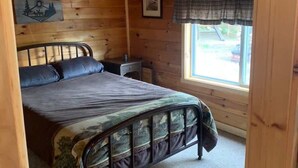 Une chambre à coucher dans un chalet en bois, avec un lit, une table de chevet, une fenêtre avec des rideaux à carreaux et un tableau accroché au mur.