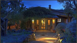 Une entrée accueillante et bien éclairée, avec un chemin de pierre menant à une porte en bois, le tout entouré d’une végétation luxuriante et d’un petit jardin.