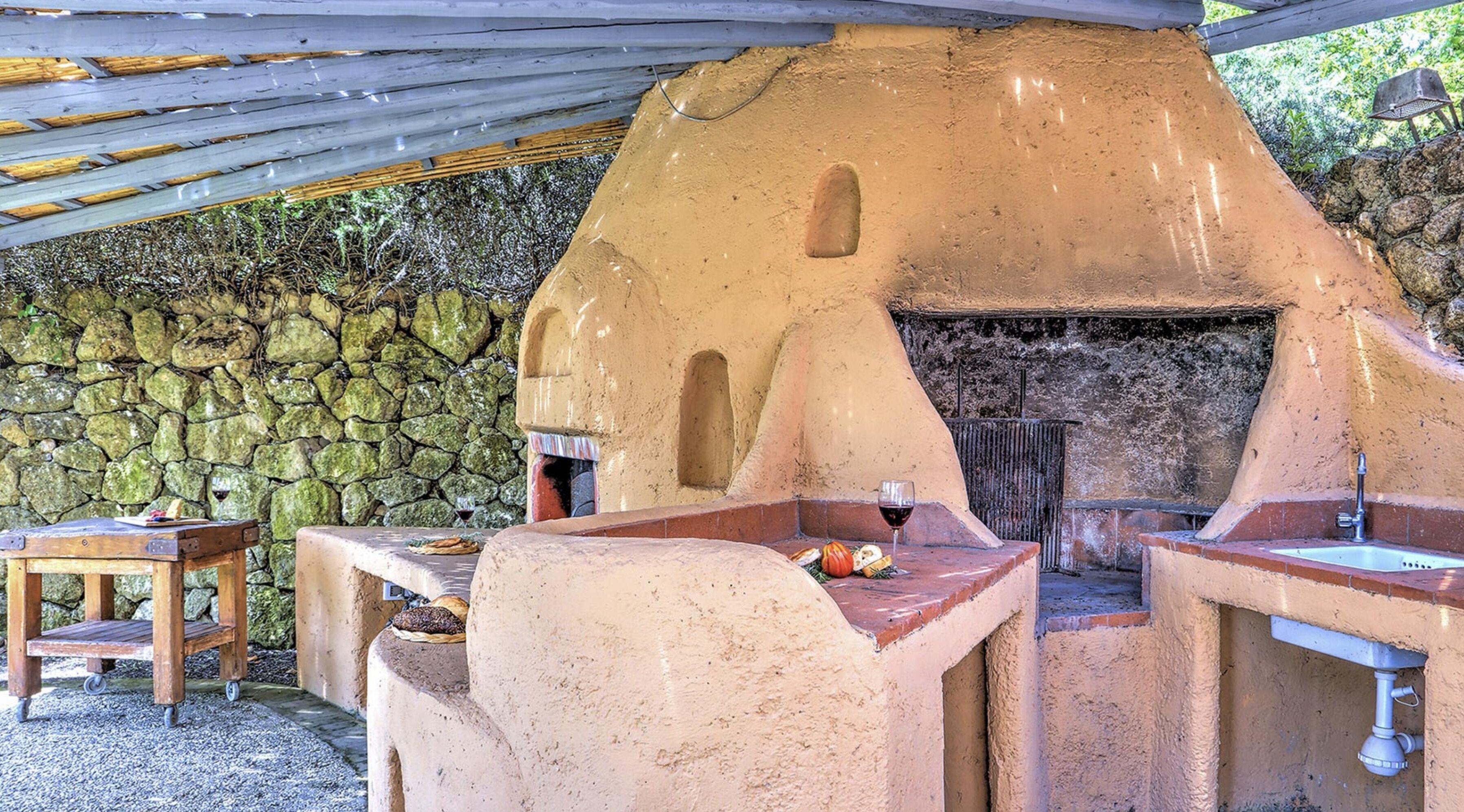 An outdoor brick oven with a stone wall backdrop, a wooden table with bread, and a metal chair.