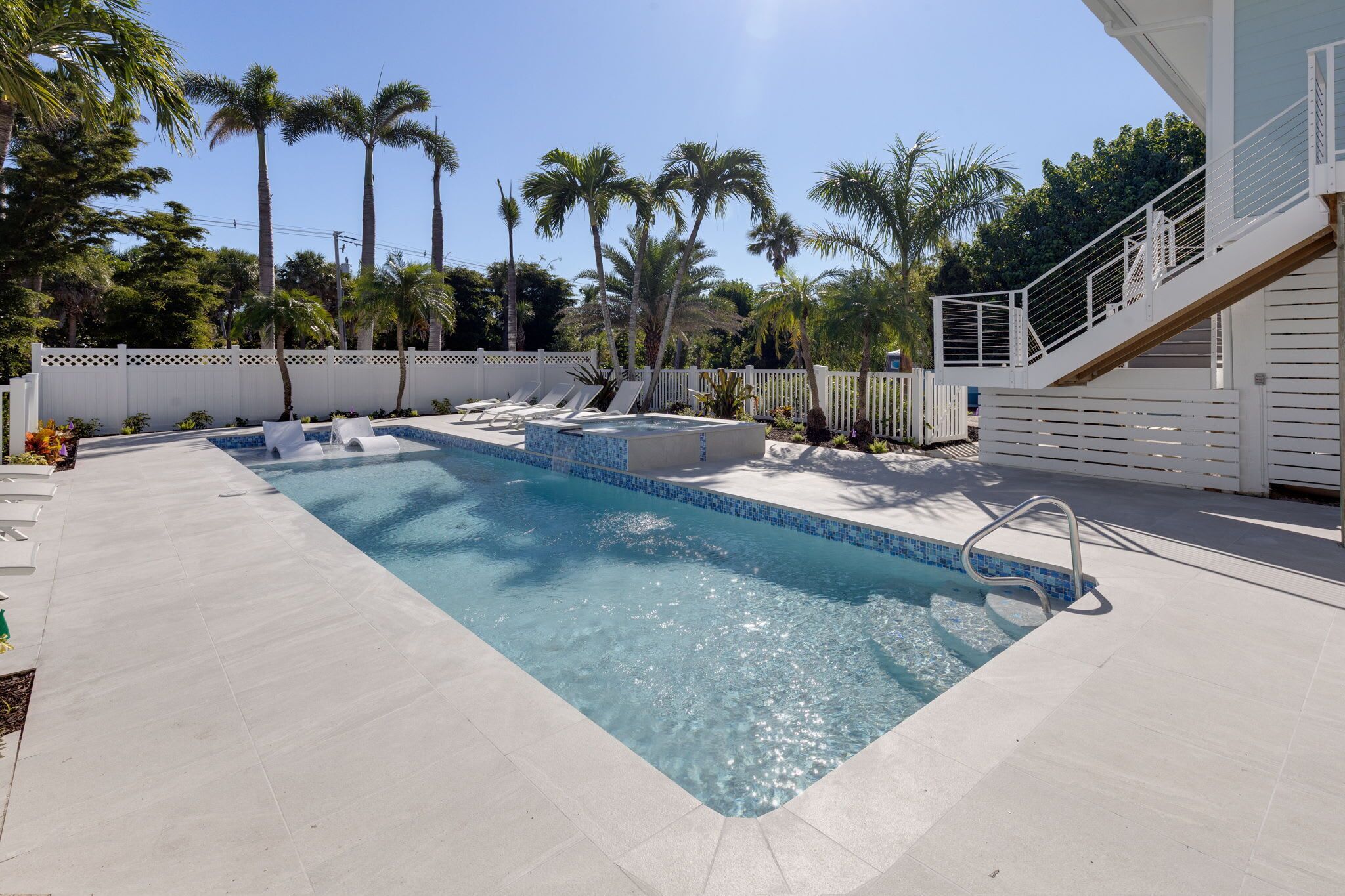 A rectangular swimming pool with a tiled edge, surrounded by a white fence and palm trees.