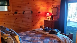 A cozy cabin room with a wooden interior, a bed with a quilt, a nightstand with a lamp, and a window with a view of trees.