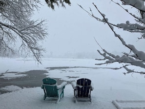 Zwei mit Schnee bedeckte Stühle blicken auf eine zugefrorene Wasserfläche, im Hintergrund kahl stehende Bäume.