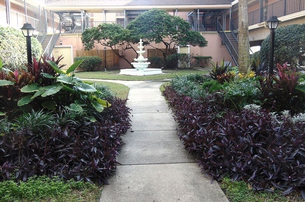 A courtyard with a central fountain, surrounded by well-maintained hedges and a pathway leading to a building with a staircase.
