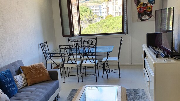 A compact living space with a sofa, a dining table with chairs, a glass-top coffee table, a TV, and a window with a view of buildings.