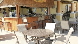 Un área de bar al aire libre con techo de paja, varios arreglos de asientos y una barra visible.