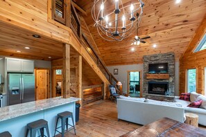 A spacious cabin interior with a kitchen, dining area, and living room, featuring wooden floors, a stone fireplace, and a large window.
