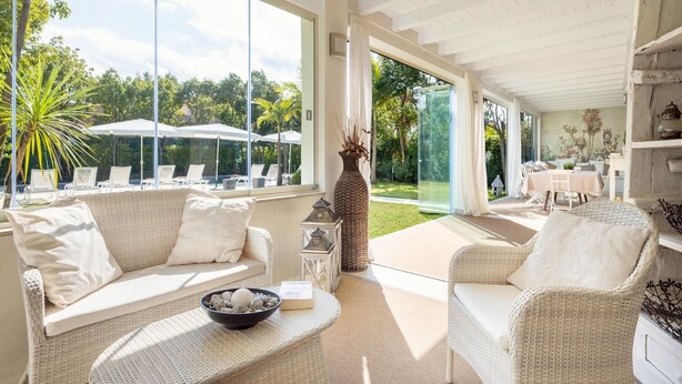 A sunlit room with wicker furniture, large windows, and a view of a garden.