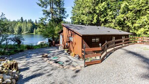 A wooden cabin with a gravel driveway, a wooden deck, and a view of a lake surrounded by trees. - Lake access cabin near trails and spring (Black Diamond)