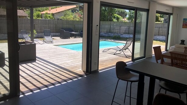Une cuisine moderne dotée d’un grand îlot central, d’un coin repas avec une table et des chaises, et d’une porte-fenêtre coulissante donnant sur une cour arrière avec une piscine et un espace salon extérieur.