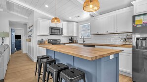 A modern kitchen with white cabinets, a wooden island with bar stools, a stainless steel refrigerator, and pendant lighting. - Sawyer's Shore Shack: Spacious, Modern Second Row Beach Home on the West End (Oak Island)