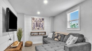 A modern living room with a grey sectional sofa, a flat-screen TV mounted on the wall, a decorative wall piece, and a window with blinds. - Sawyer's Shore Shack: Spacious, Modern Second Row Beach Home on the West End (Oak Island)