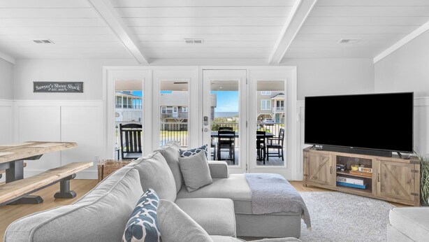 A living room with a grey sectional sofa, a wooden coffee table, a flat-screen TV on a wooden stand, and a view of the beach through glass doors. - Sawyer's Shore Shack: Spacious, Modern Second Row Beach Home on the West End (Oak Island)