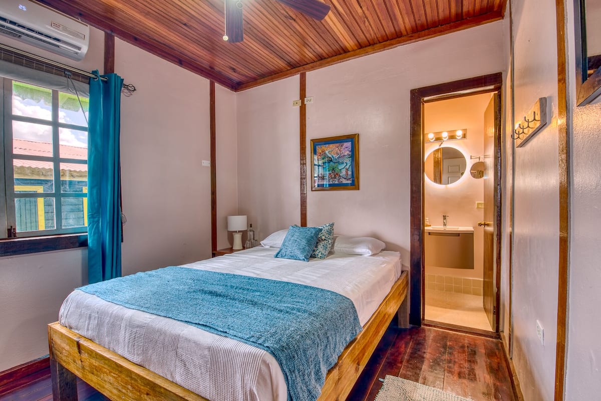 A bedroom with a bed, a window with curtains, a painting on the wall, and a bathroom visible through an open door.