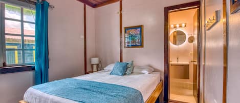 Quarto com cama, janela com cortinas, quadro na parede e banheiro visível por uma porta aberta.