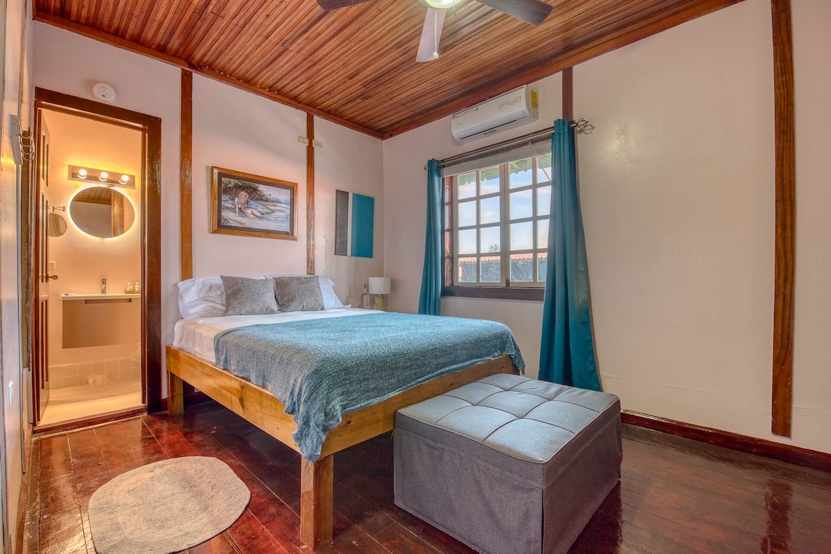 A bedroom with a wooden bed, a small bedside table, a window with curtains, and a ceiling fan.