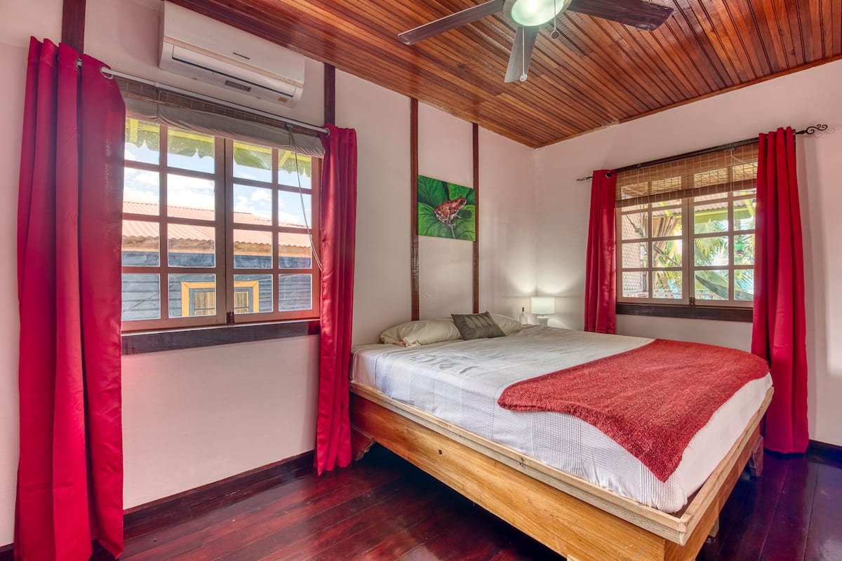 A bedroom with a wooden bed, red curtains, a ceiling fan, and a window with a view of buildings.