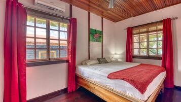 Un dormitorio con cama de madera, cortinas rojas, ventilador de techo y ventana con vistas a edificios.