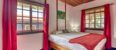 Une chambre avec un lit en bois, des rideaux rouges, un ventilateur de plafond et une fenêtre donnant sur des immeubles.