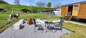 Un espace extérieur aménagé dans un style rustique, avec une table et des chaises, une structure en bois dotée d’une porte ouverte, et un espace clos où se trouve un cheval.