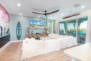 A modern living room with a white sectional sofa, a ceiling fan, a surfboard decoration, and a beach-themed painting.